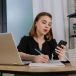 Business professional comparing hosted phone system vs traditional PBX costs on mobile device while taking notes in Dallas Fort Worth office