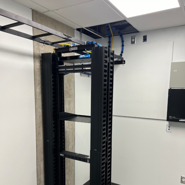 Fully installed network rack with organized blue and yellow cable runs entering through ceiling cable tray, adjacent to access control panel